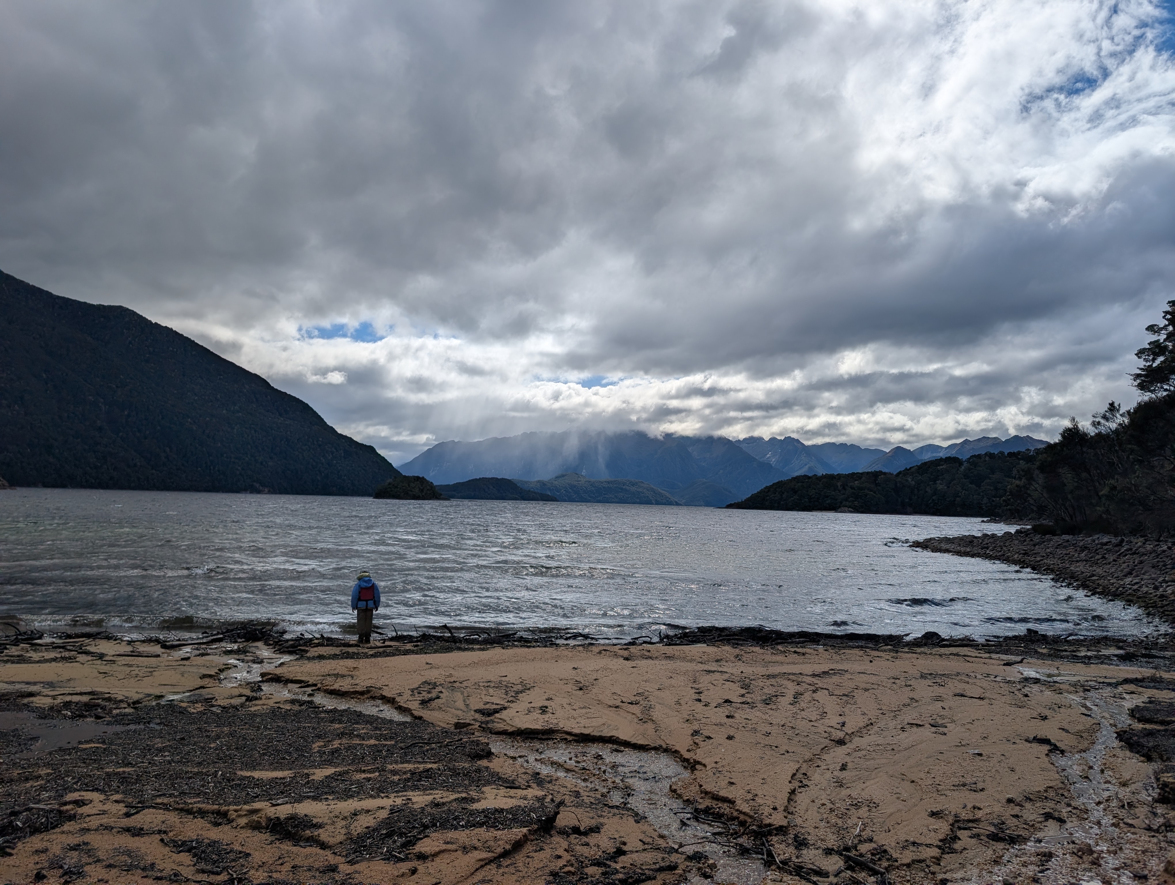 A person standing on a sandy shore looking out at a calm lake surrounded by mountains under a cloudy sky.