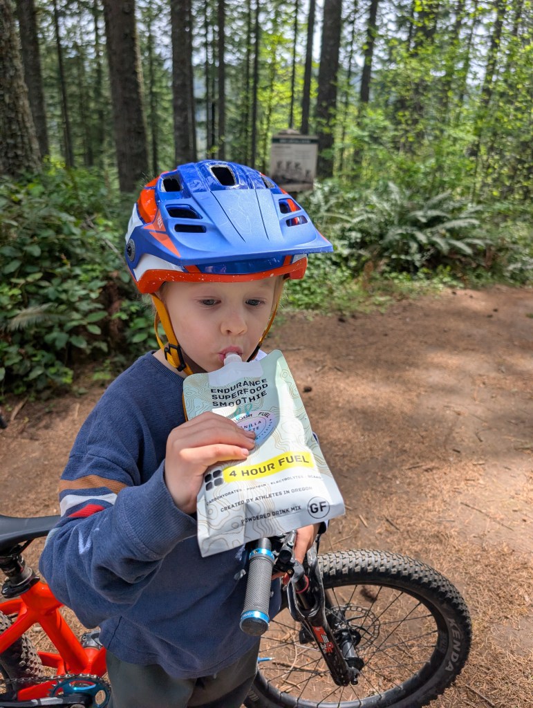 A young child wearing a blue and orange bike helmet is standing on a forest trail, holding a pouch labeled 'Endurance Superfood Smoothie 4 Hour Fuel' while sipping from it. A red bicycle is partially visible next to the child amidst tall trees and greenery.