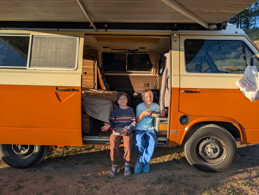 Two children sitting on the steps of a vintage orange camper van, enjoying snacks. The interior of the van is visible, showcasing a cozy setup.